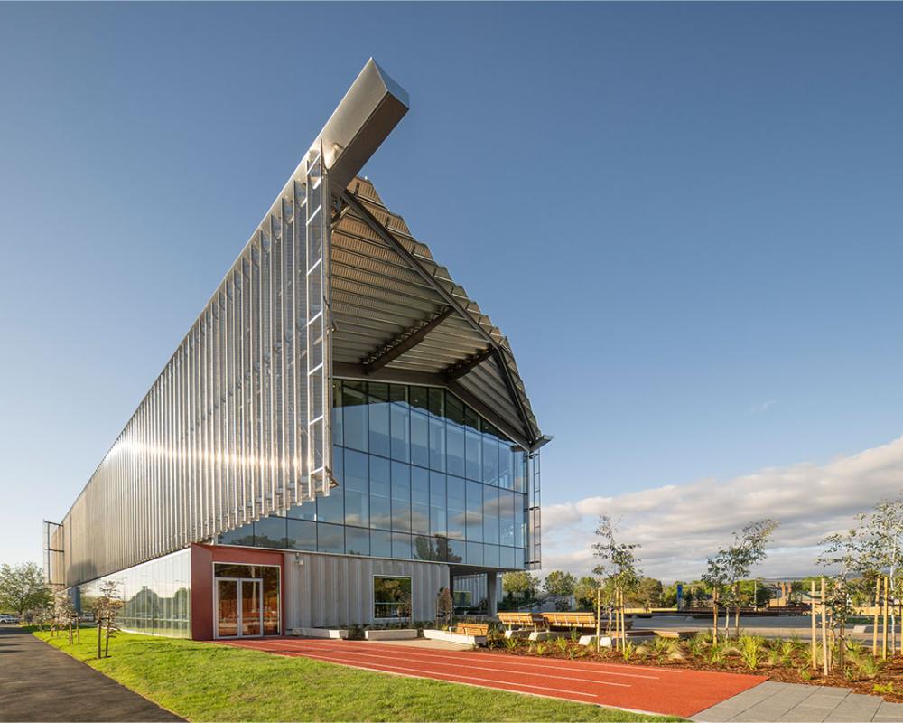 Modern building with a steel facade, large glass windows, and a unique angular roof, reflecting sunlight.