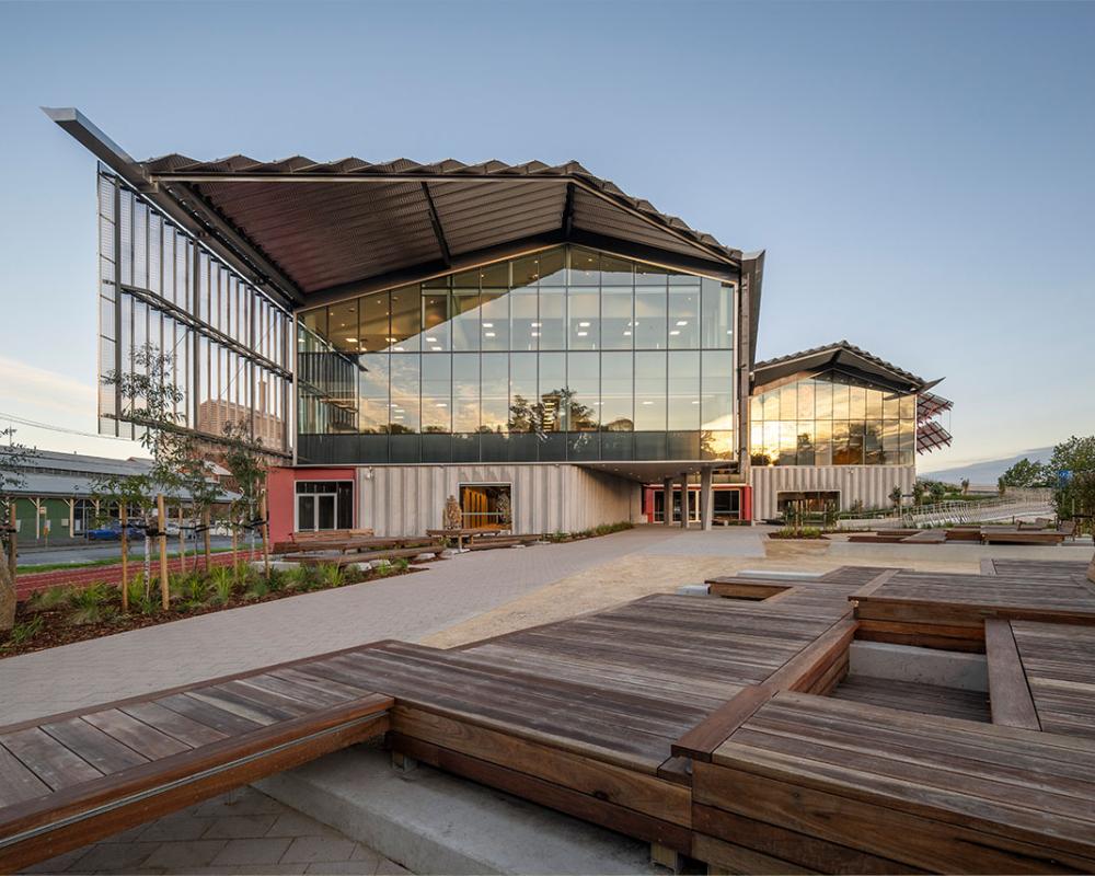 Modern architectural building with large glass windows, an angular metal roof, and an outdoor seating area with wooden benches, surrounded by landscaped greenery.