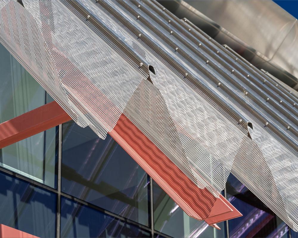 Close-up of perforated metal sunshade with red steel supports on a modern glass building façade.