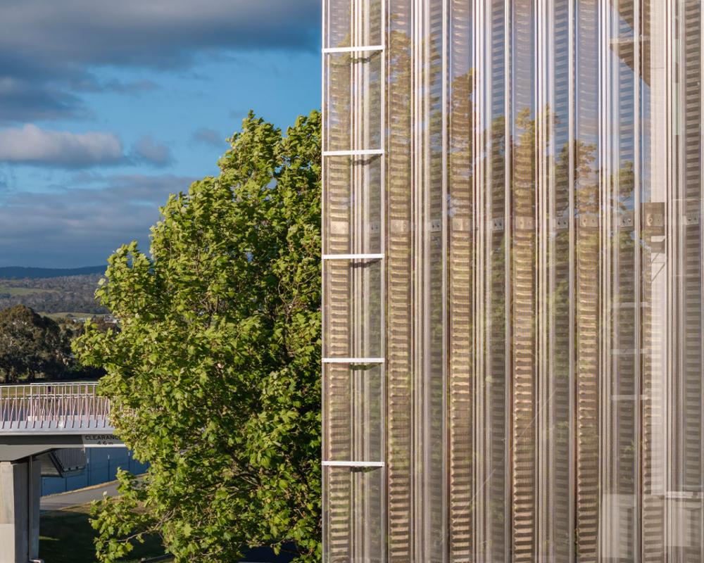 Close-up of a reflective metal façade with vertical louvers, mirroring trees and sky in the background.