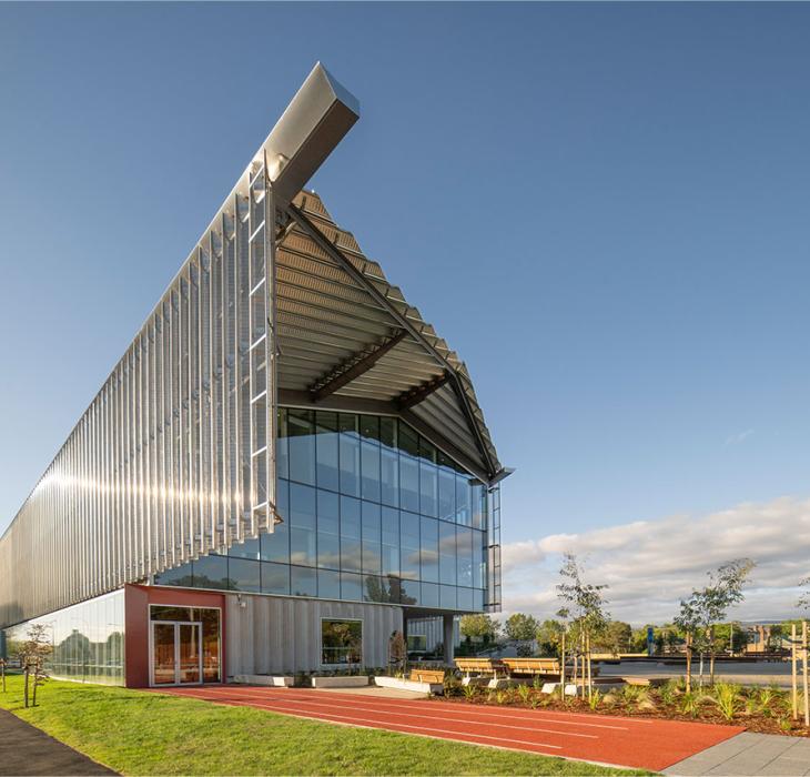 Modern building with a steel facade, large glass windows, and a unique angular roof, reflecting sunlight.