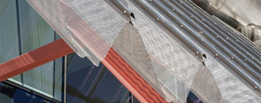 Close-up of perforated metal sunshade with red steel supports on a modern glass building façade.